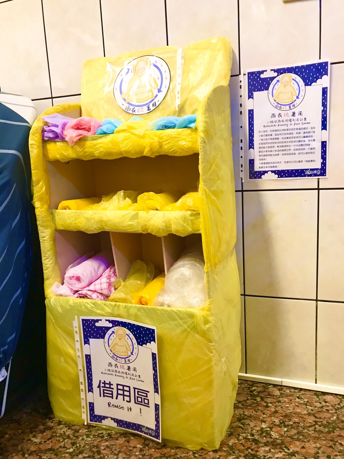 「雨衣琉著用」雨衣陳列架。由便利商店閒置的展示架再利用而成,由於材質是紙類,因此外表還以廢棄輕便型雨衣包覆,避免在雨天受潮毀損。(陳汯葰/攝) 「雨衣琉著用」雨衣陳列架。由便利商店閒置的展示架再利用而成,由於材質是紙類,因此外表還以廢棄輕便型雨衣包覆,避免在雨天受潮毀損。(陳汯葰/攝)